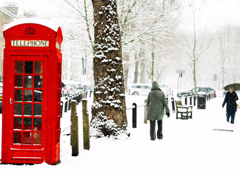 A London street covered in snow with people walking and a red telephone box.